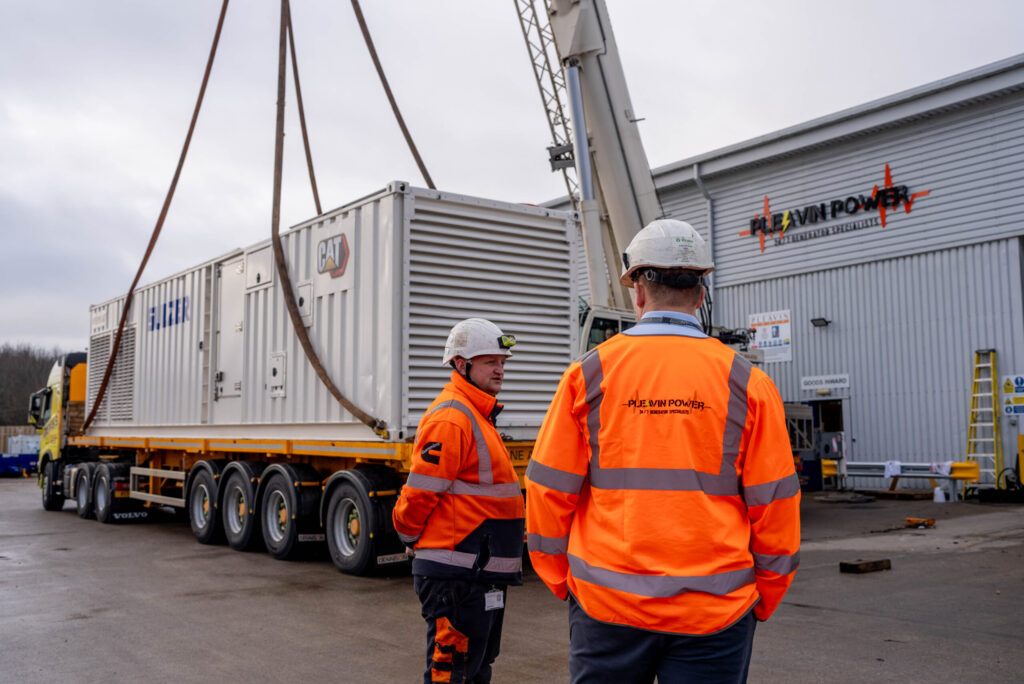 Two engineers in hi-vis gear and safety helmets oversee a crane moving a geneator unit.
